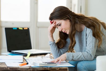 woman sits at table surrounded by paperwork. There is a laptop and she is using a calculator. She has a worried expression.